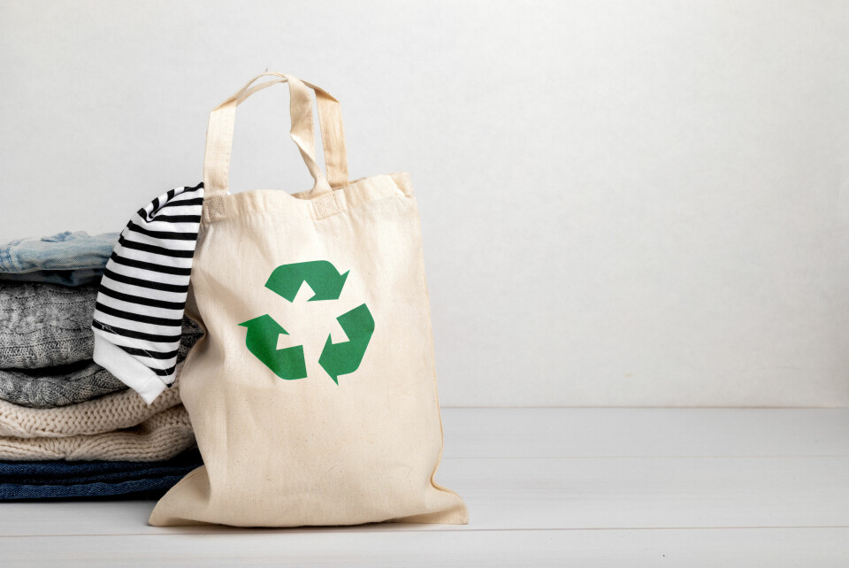 A photo of a fabric bag with the recycling logo on it, filled with clothes, next to a pile of folded clothes
