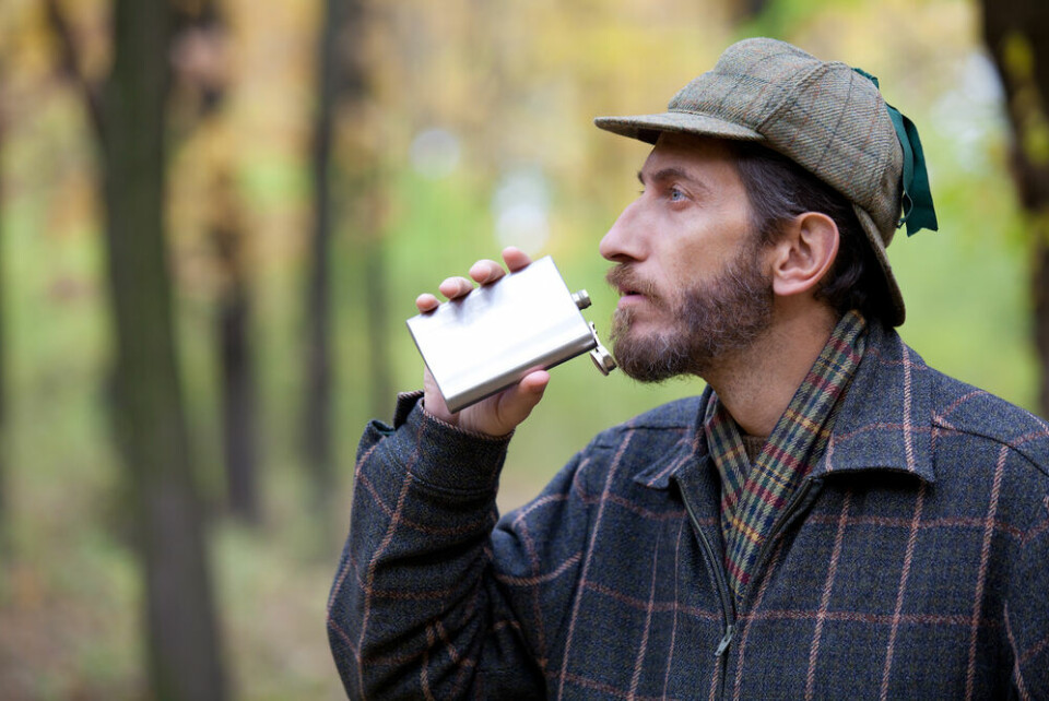 A photo of a man wearing a hunting hat, taking a drink from a hip flask while outdoors