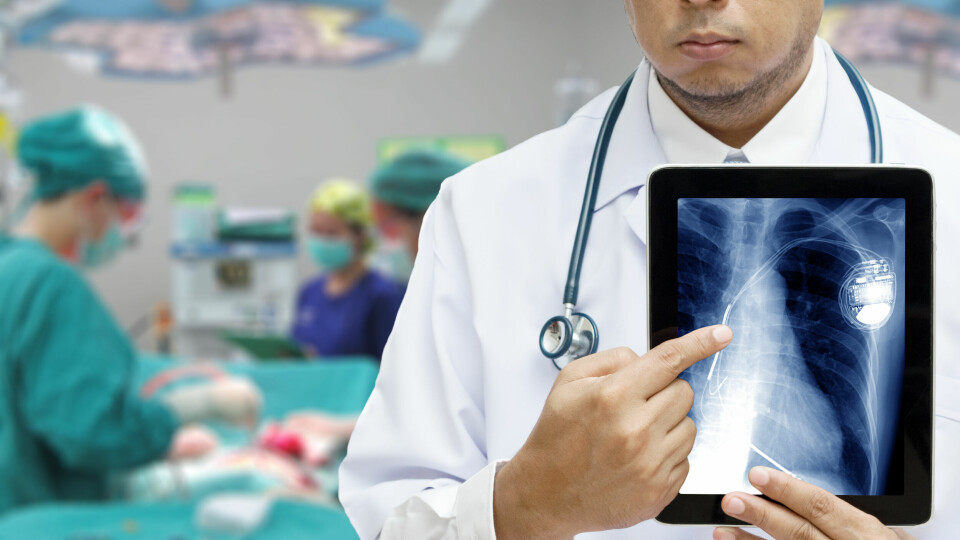A photo of a doctor holding a tablet with an X-ray of a pacemaker in the foreground with surgery happening in the background
