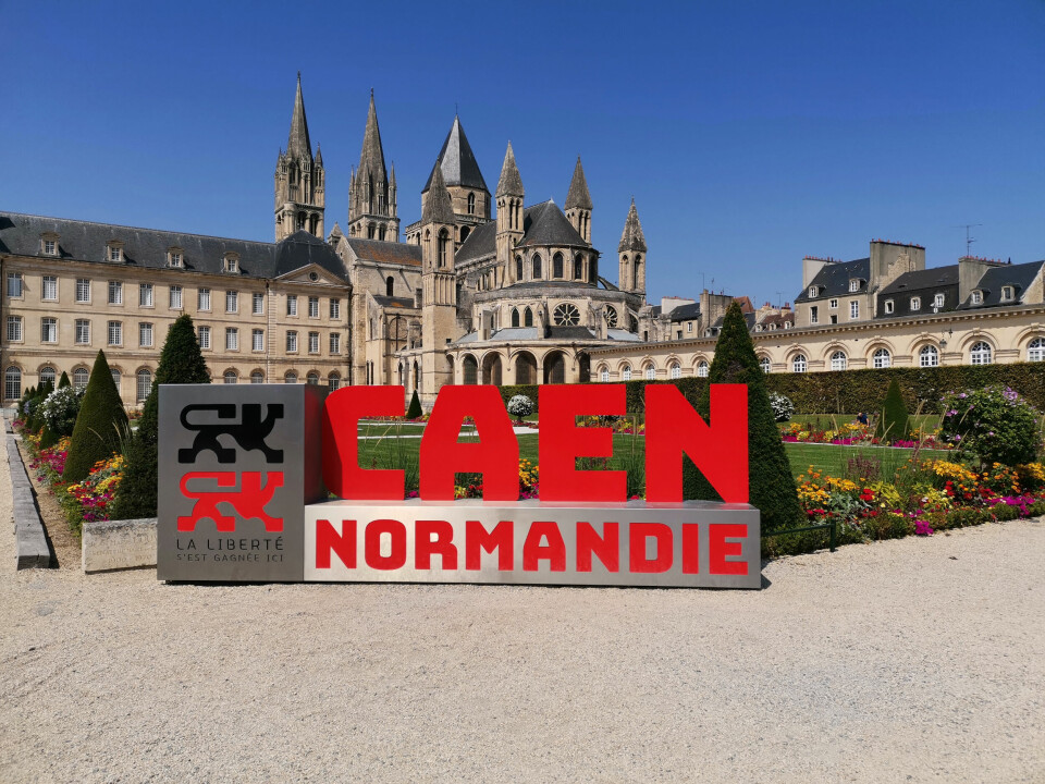 A metal sign reading CAEN, Normandie, in front of Caen cathedral