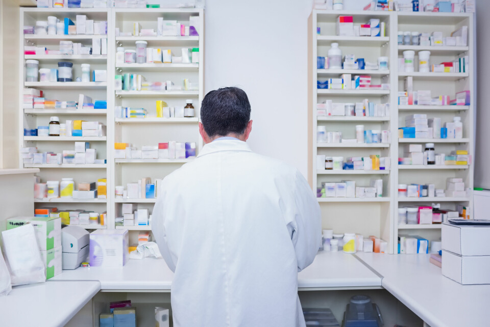 A pharmacist wearing a white coat stands with his back to the camera surrounded by medicine on shelves
