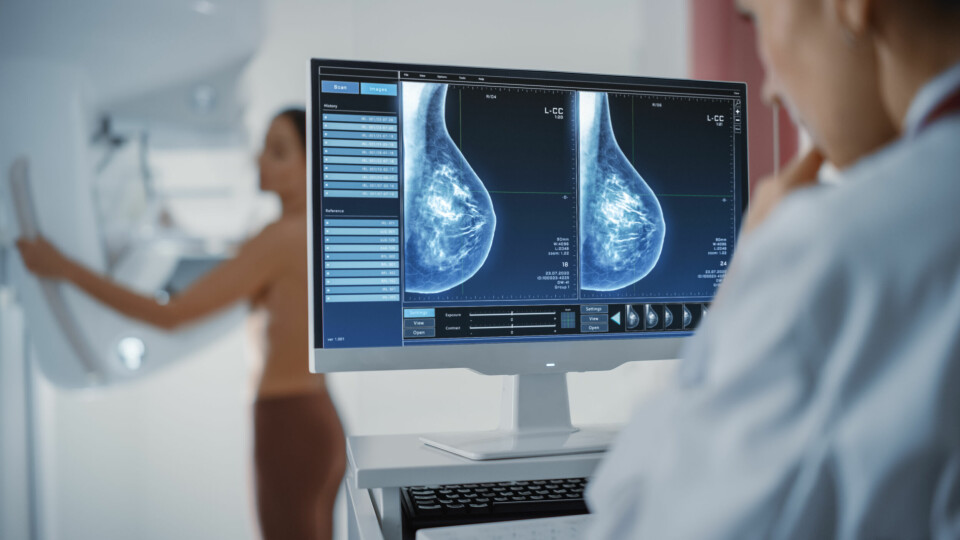 A photo of a radiologist looking at a mammogram screening with a woman being examined in the background