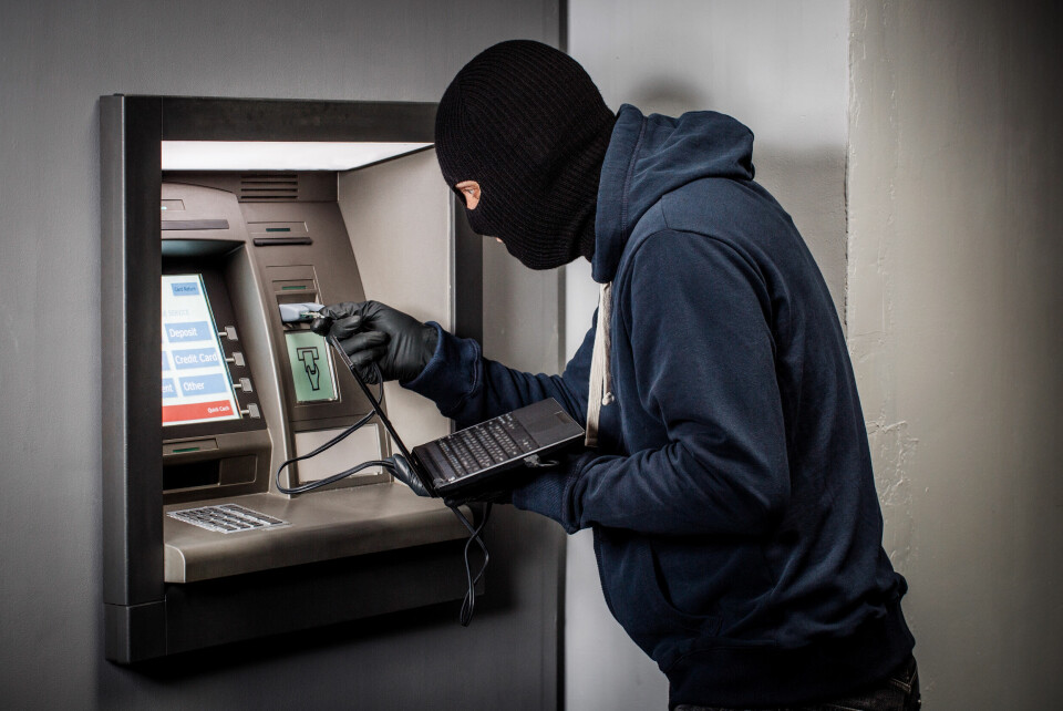 A hacker wearing a balaclava using a laptop to steal money from an ATM