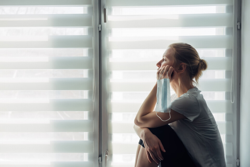 exebiche / Shutterstock A woman holding a mask sits by a window looking fed up