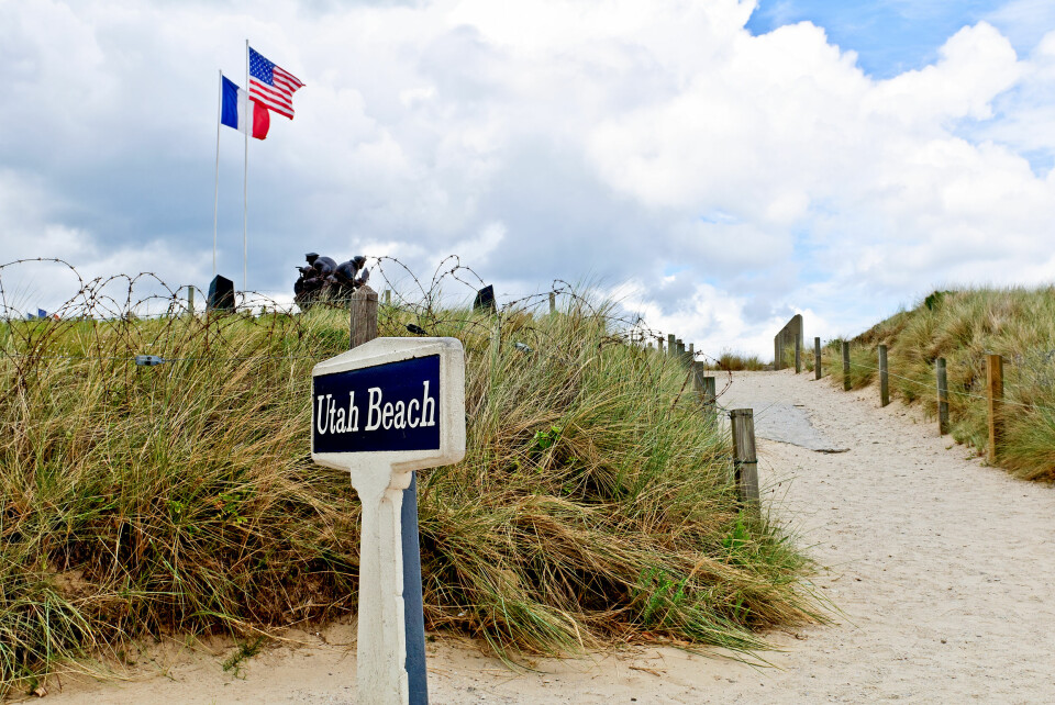 A sign in the sand at the entrance to Utah Beach, one of the sites of the D-Day landings, with US and French flags flying at a memorial in the background