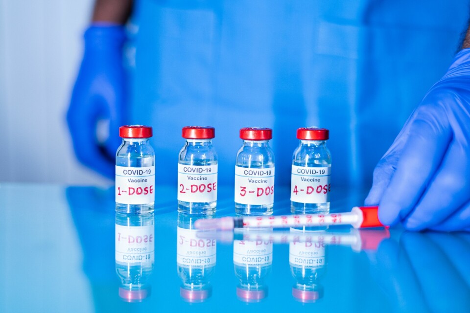 A nurse wearing gloves stands behind four vials of Covid vaccine, labelled 1 to 4, and a syringe