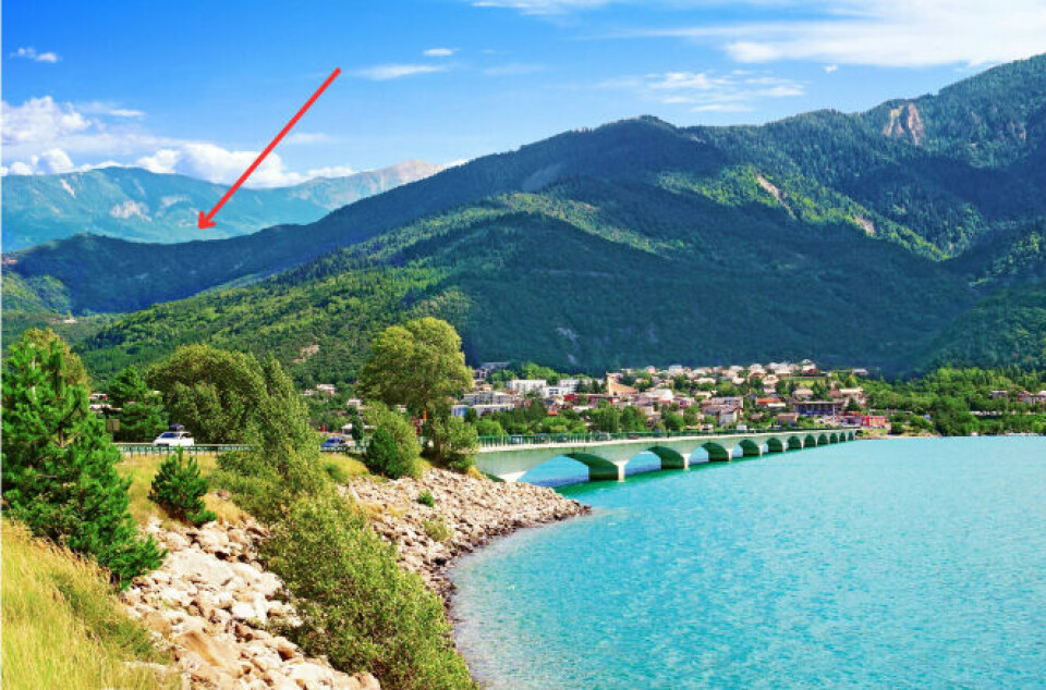 The bridge of Savines-le-lac in the French Alps, over a idyllic blue lake. An arrow points over a ridge in the distance.