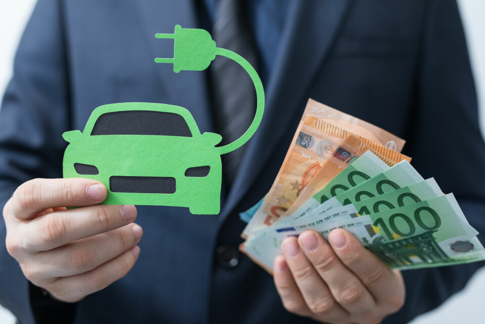 A photo concept of electric car loans, with a man holding a paper cut-out of an electric car with a plug and holding euro notes in the other hand