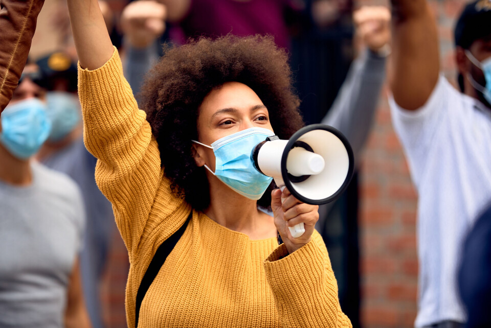 Drazen Zigic / Shutterstock A woman wearing a mask and holding a megaphone raises her fist in the air during a protest with other people wearing masks behind her