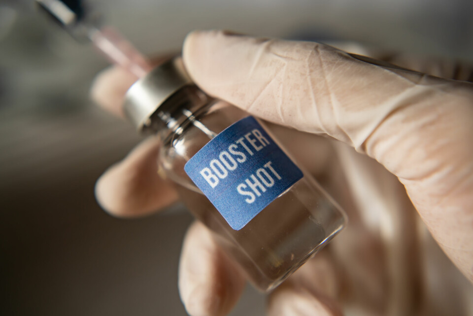 A gloved hand holding a vial labelled ‘booster shot’ with a syringe preparing to use it