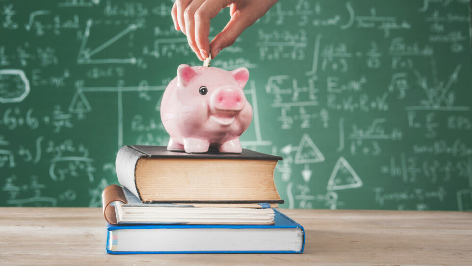 A woman puts coins into a piggybank on top of books, with a blackboard behind it. Back to school financial grant is on the way to 3 million French homes