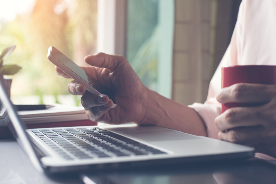 An image of a man's hands holding a phone and typing something on a laptop