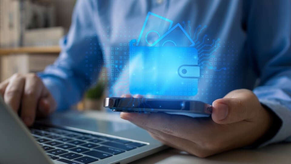 A man sitting at a computer holding a phone with a visual of a wallet and money to show bank transfers