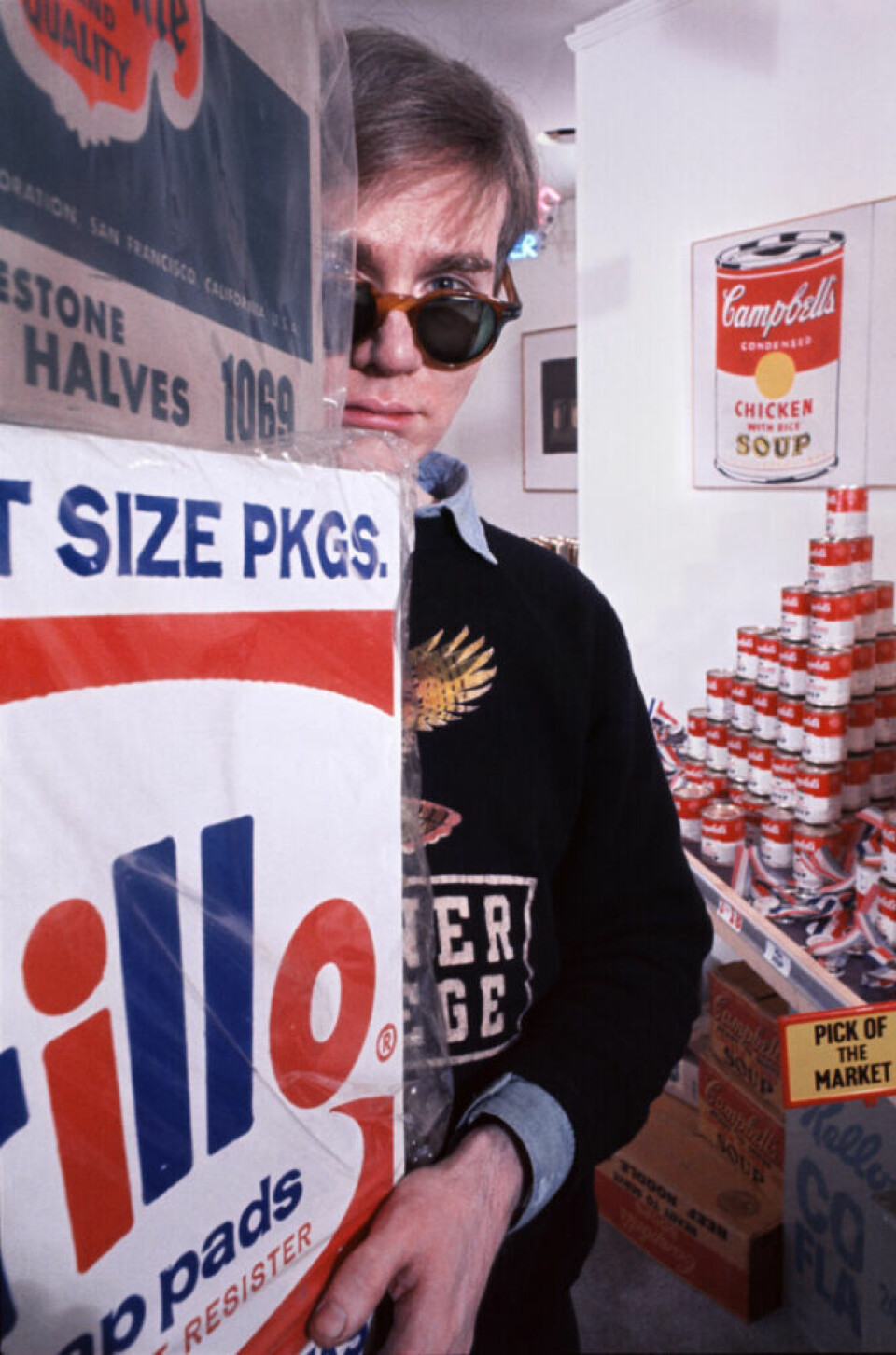 Andy Warhol wearing sunglasses and standing behind a sign at his The American Supermarket exhibition