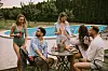 A group of young adults drinking and eating a table near a swimming pool
