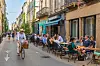 Man on a bike cycling through a street in Bordeaux with people eating and drinking outside restaurants