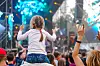 A young girl sits on someone's shoulders in a festival crowd
