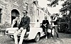 Four men with instruments pose around a classic white car outside an old stone building.