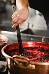 Hand stirring a dark berry mixture in a large copper pot in a kitchen.