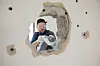 Worker using a power drill to demolish an interior wall seen through a hole