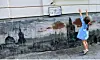 Young girl reaching up beside a painted cityscape mural on a wall
