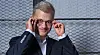 Person in a dark suit adjusting round glasses before a wire mesh background