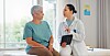 Doctor holding a tablet while talking with an older patient in a clinic room.
