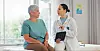 Doctor holding a tablet while talking with an older patient in a clinic room.