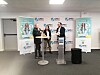 Three people speak at podiums in front of SMD3 banners at an indoor event.