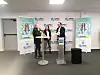 Three people speak at podiums in front of SMD3 banners at an indoor event.