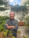 Smiling man stands with folded arms in a garden courtyard with plants and benches behind him.
