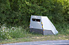 A roadside speed camera enclosure beside a rural road, partly obscured by grass and bushes.