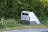 A roadside speed camera enclosure beside a rural road, partly obscured by grass and bushes.