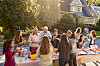 Neighbours stand and sit around a long table outdoors, talking and eating at a block gathering.