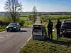 A damaged roadside speed camera stands near a rural road, with two police officers and a vehicle nearby.