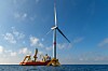 Floating offshore wind installation with cranes and a turbine on a barge at sea under a blue sky.