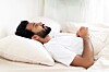 Indian man lying in bed, awake and looking distressed, with white pillows and bedding.