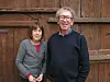 Two people standing in front of a wooden wall and door outdoors.