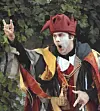 Man in a medieval-style costume gesturing with raised hand in front of green foliage.