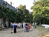 People standing and chatting on a gravel courtyard beside trees and white chairs at an outdoor gathering.