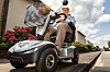 Elderly person driving an electric mobility scooter beside a roadside verge.