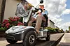 Elderly person driving an electric mobility scooter beside a roadside verge.