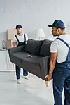 Two movers in uniform carry a grey sofa in a bright room.