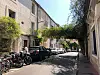 Narrow street with parked cars, motorbikes, and stone buildings under trees.
