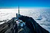 Mountain summit with antenna and buildings above clouds under a blue sky.