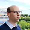 Man wearing glasses standing on a balcony with buildings, trees and cloudy sky behind him.