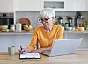 Senior woman sitting at a kitchen table using a laptop and writing in a notebook.