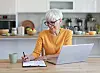 Senior woman sitting at a kitchen table using a laptop and writing in a notebook.