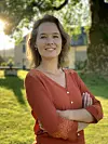 Smiling woman standing outdoors with arms crossed in a sunny garden.