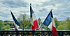 French flags waving above a bridge railing with Paris buildings in the distance.